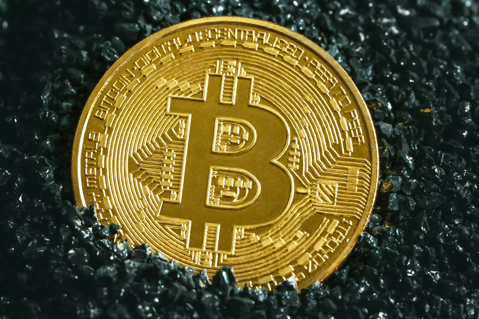 A close-up of a golden Bitcoin coin placed on a bed of dark, glistening stones, symbolizing the value and prominence of cryptocurrency in modern finance.