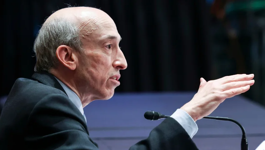 Gary Gensler, président de la SEC, prenant la parole lors d'un événement public, faisant un geste de la main tout en étant assis devant un microphone